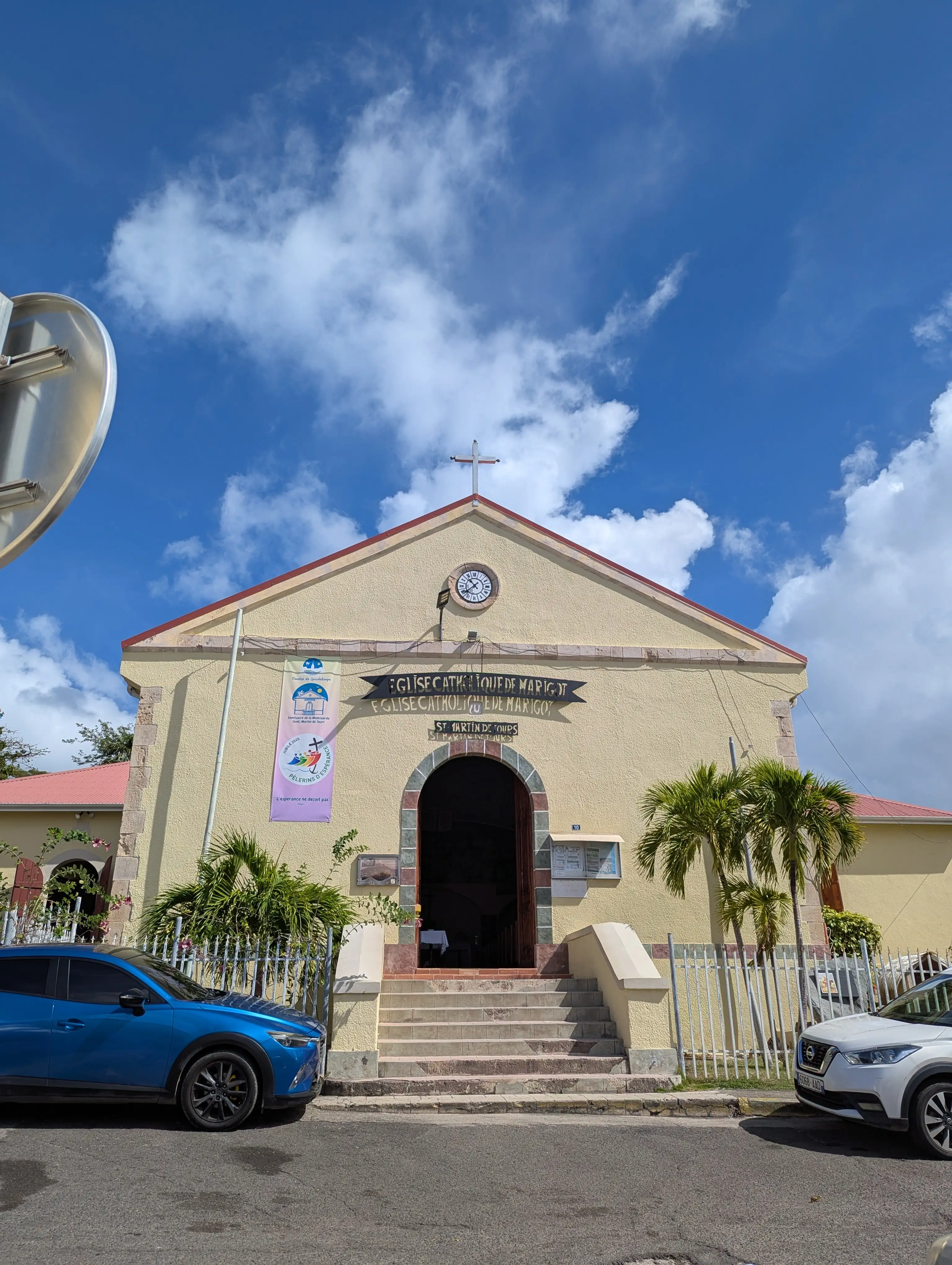The cream-colored Catholic church in Marigot, Saint-Martin, seen head-on from the street with its bell tower and red roof under a partly cloudy sky
