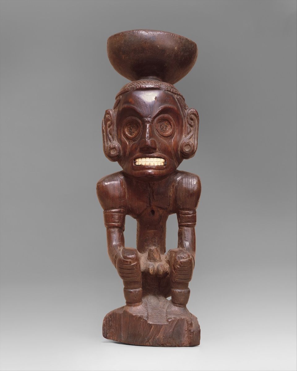 A carved stone figure of a crouching deity with a broad, open-mouthed face and arms braced against its knees.