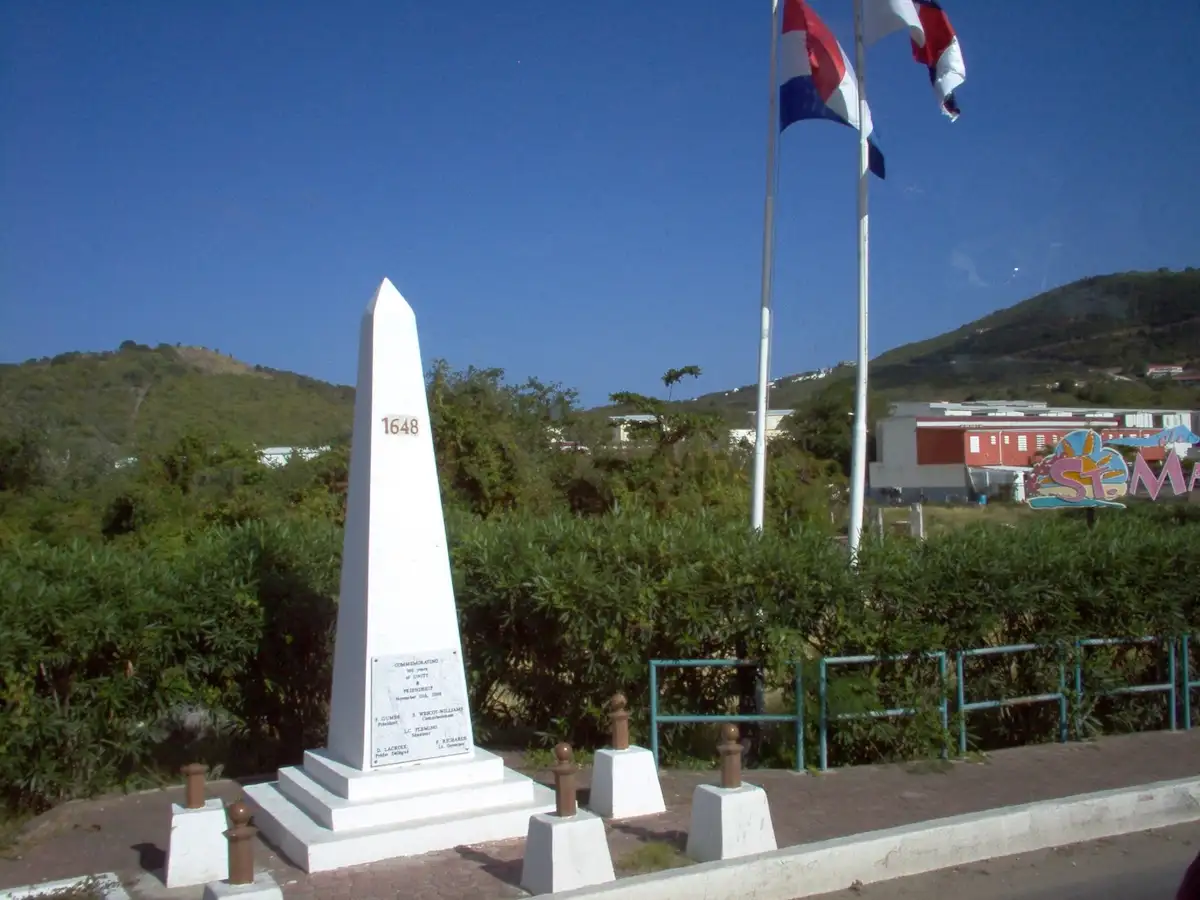 A white stone obelisk beside a road marking the boundary between the French and Dutch sides of the island, with flags flying and green hills behind