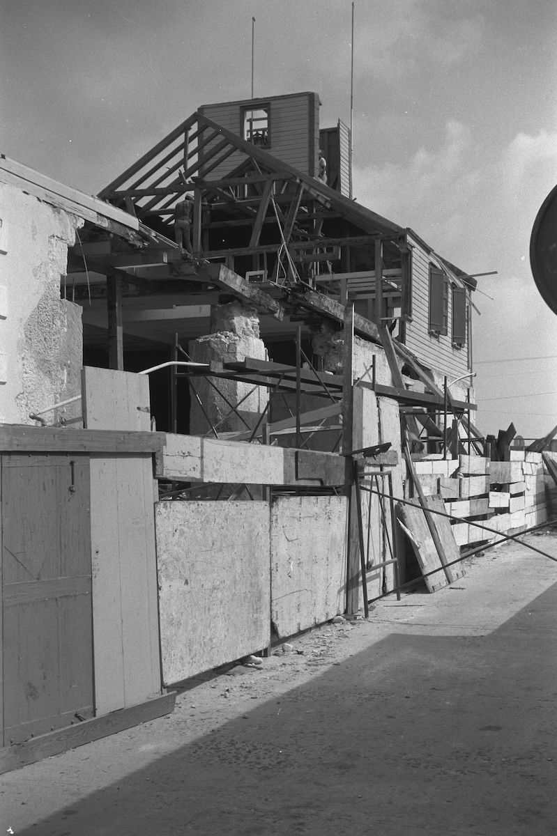 The rear of the Philipsburg Courthouse stripped back to its timber frame during restoration, with stacked stone and boards in the yard and men working on the exposed roof structure.
