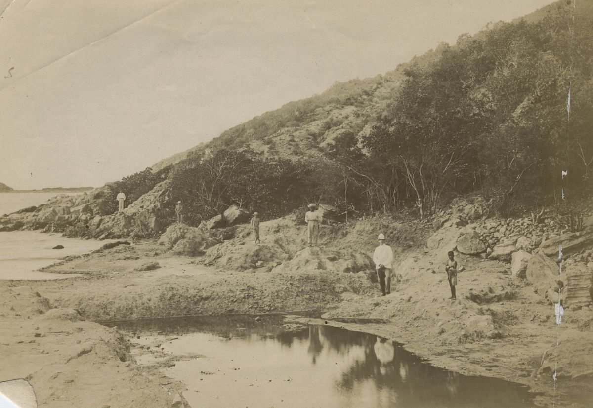 Several men stand at the edge of a worked salt pond, some in white colonial dress and pith helmets, beside a cut channel in the pan.