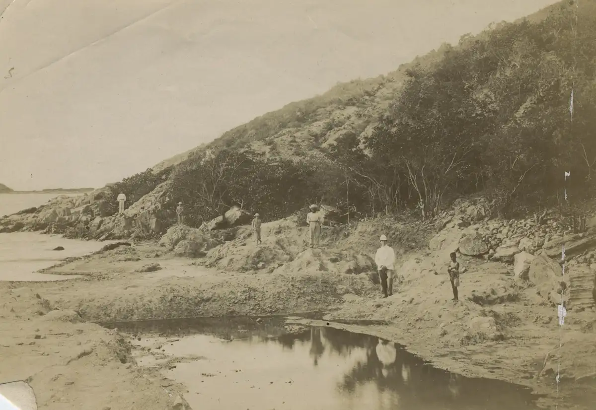 Several men stand at the edge of a worked salt pond, some in white colonial dress and pith helmets, beside a cut channel in the pan