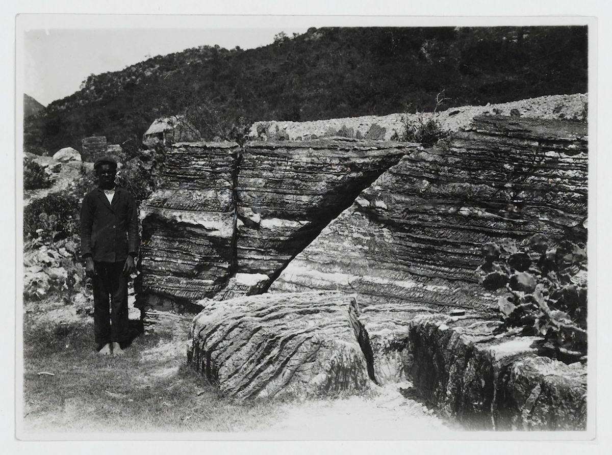 A man in a jacket and cap stands barefoot beside a large layered rock formation at the edge of a salt pan.