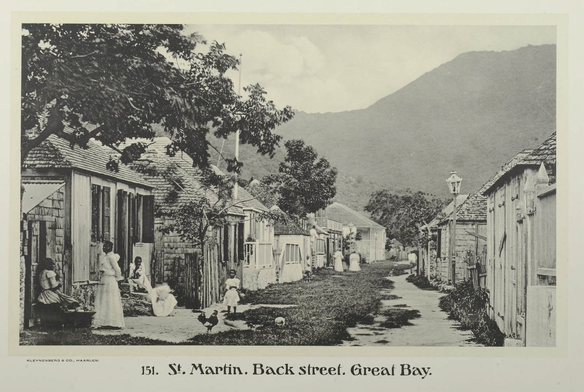 A printed photographic plate captioned St. Martin, Back street, Great Bay, showing a grassy lane between wooden shingled houses, with women in white dresses and children standing in the road, chickens in the foreground and a green hillside beyond.