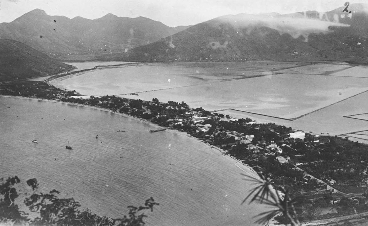A view from a hillside over Great Bay, showing the low rooftops of Philipsburg strung along a narrow sandbar between the open sea and the geometric rectangles of the salt pans behind.