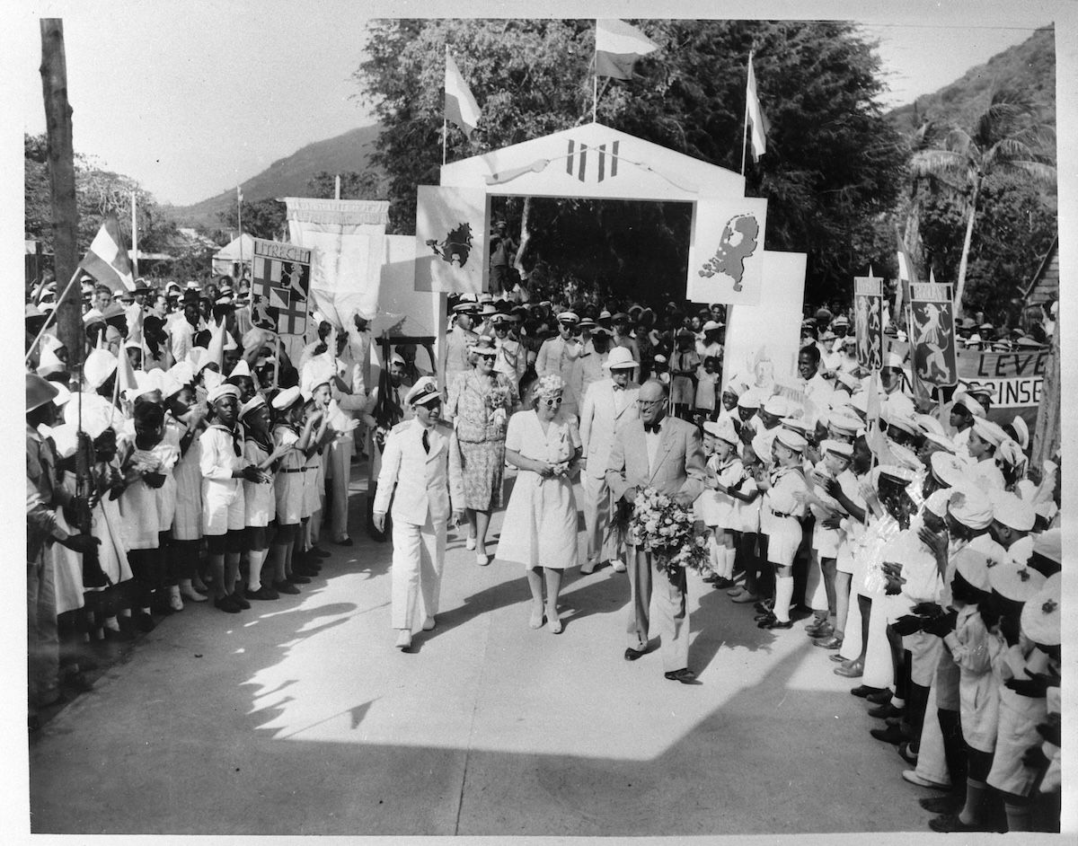 Princess Juliana in a white dress and hat walks along a path lined on both sides by hundreds of children in white uniforms waving flags, passing beneath a decorated ceremonial arch hung with provincial coats of arms.