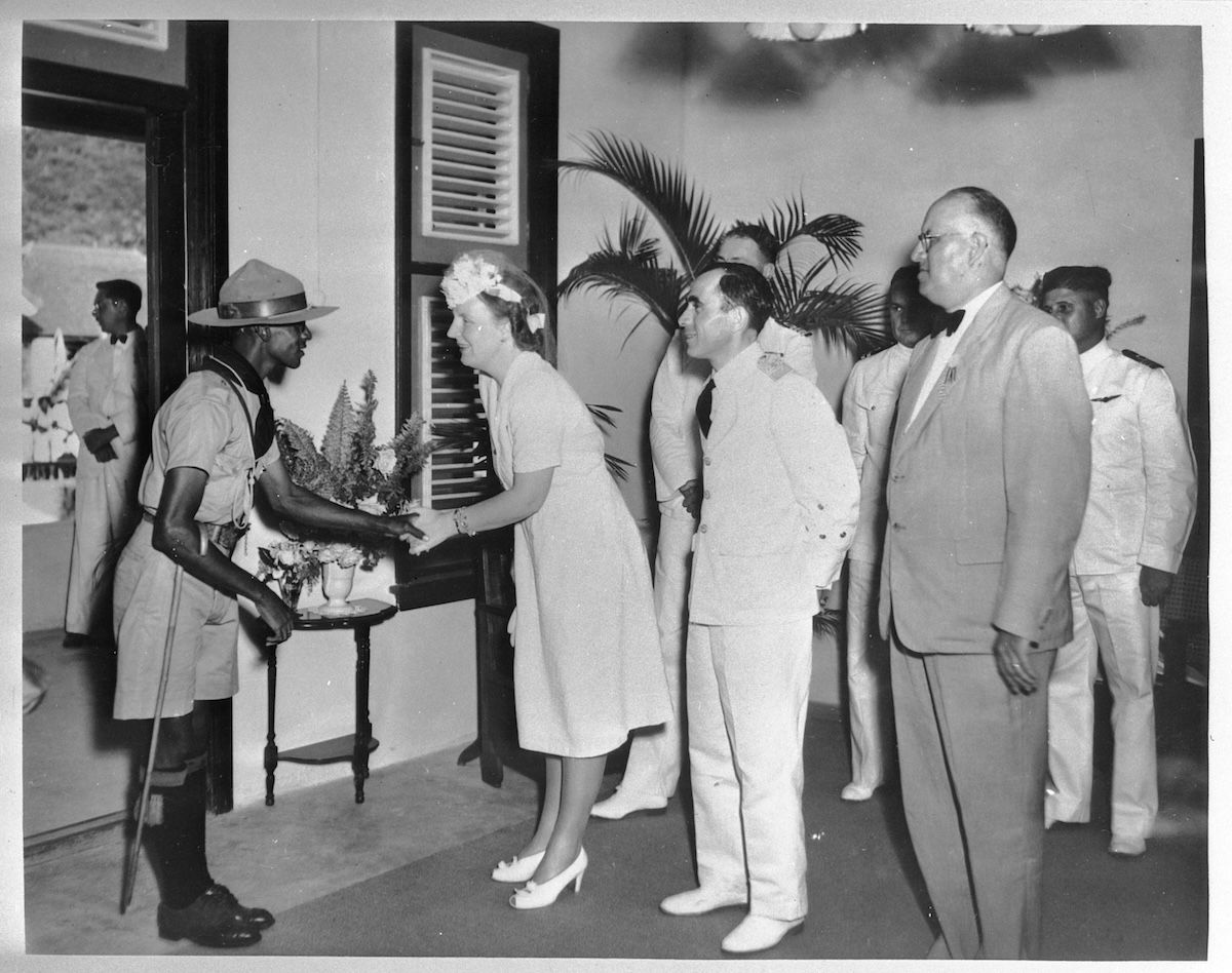 Princess Juliana shakes hands with a uniformed man in a wide-brimmed hat during a receiving line indoors, watched by officials in white suits.
