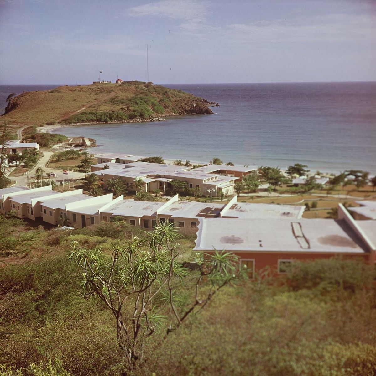 A colour photograph looking down from a hillside at a low white resort complex spread along a curved beach, with a green headland and open sea beyond and scrub in the foreground.
