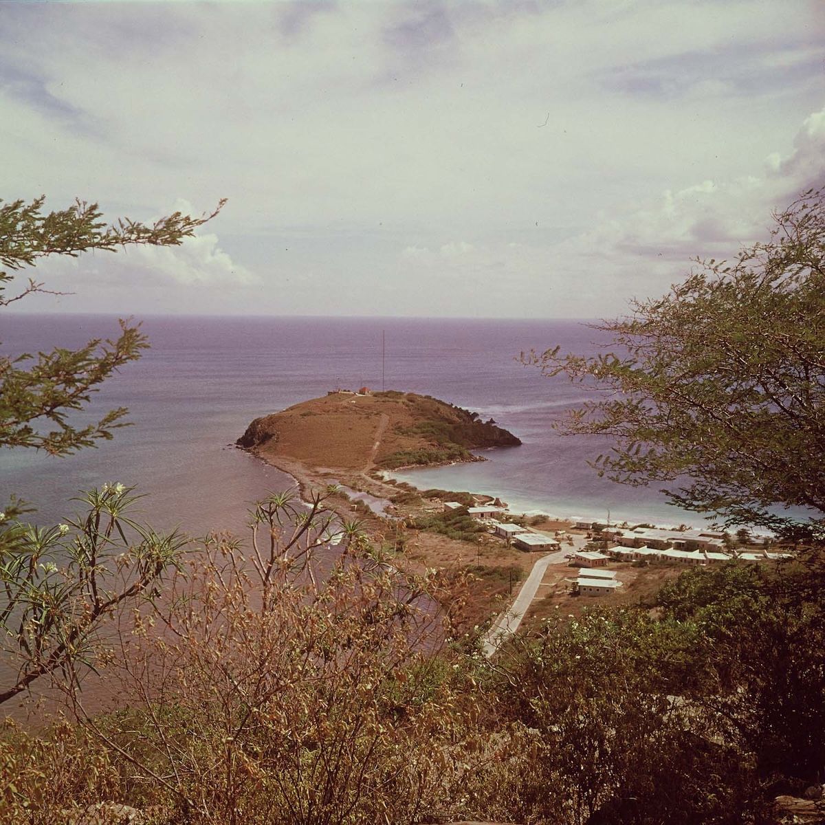 A colour view from a hillside of the peninsula carrying Fort Amsterdam, with the Little Bay Hotel buildings along the shore to the right and open sea beyond.