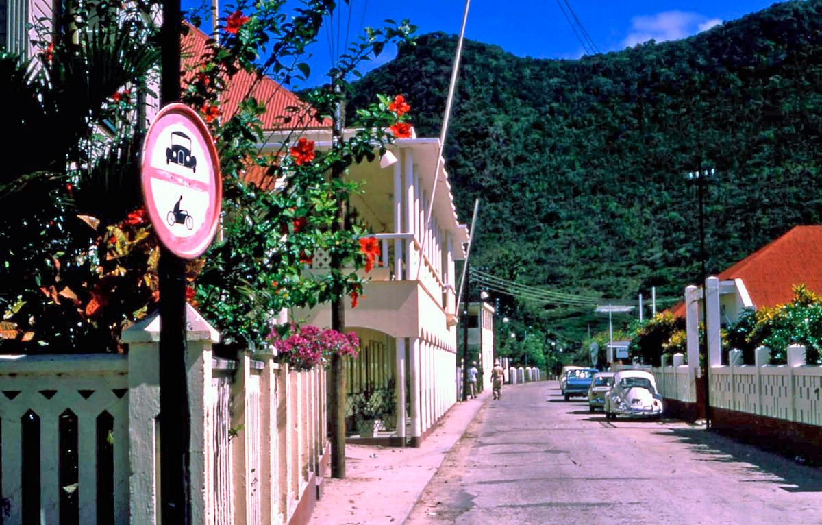 A quiet Philipsburg street with a two-storey white building with balconies, red hibiscus and bougainvillea in flower, a no-motor-vehicles sign, and a few parked cars under a green hillside.