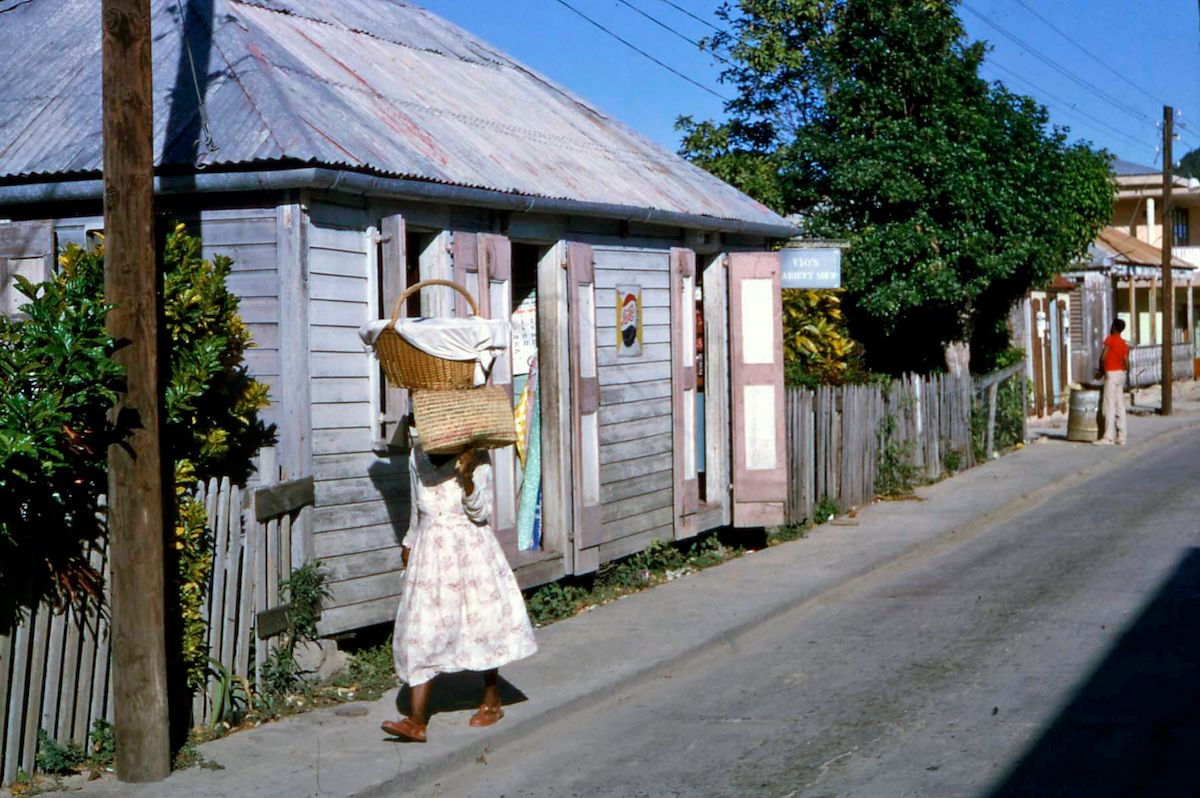 A woman in a patterned dress carries a large basket and a covered tray balanced on her head as she walks along a street past a weathered wooden shop with open shutters.