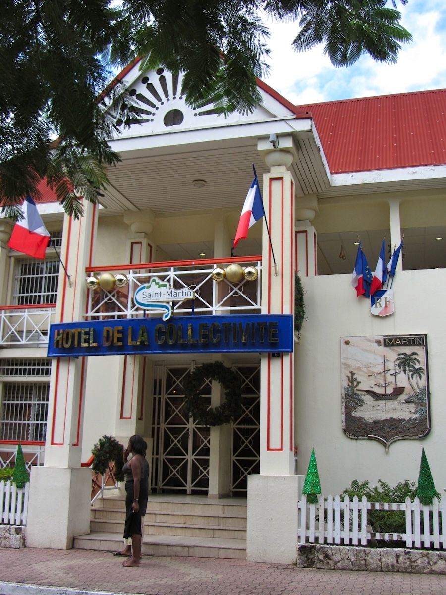 The entrance of the Hôtel de la Collectivité in Marigot, with French tricolour flags, a Saint-Martin collectivity sign over the door, an RF plaque and a carved coat of arms on the wall.