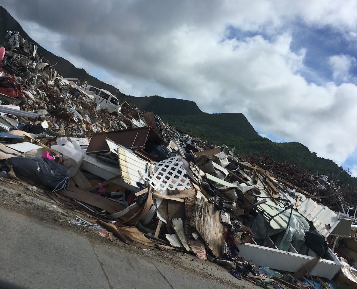 A long mound of hurricane debris piled beside a road, made up of broken timber, roofing, furniture and a wrecked van, with green hills and clouds behind.