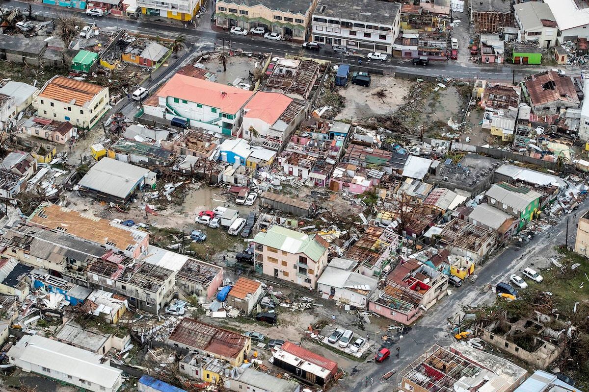 An aerial photograph of a Sint Maarten neighbourhood after Hurricane Irma, with roofs torn off dozens of houses, debris scattered across yards and streets, and damaged vehicles among the wreckage.