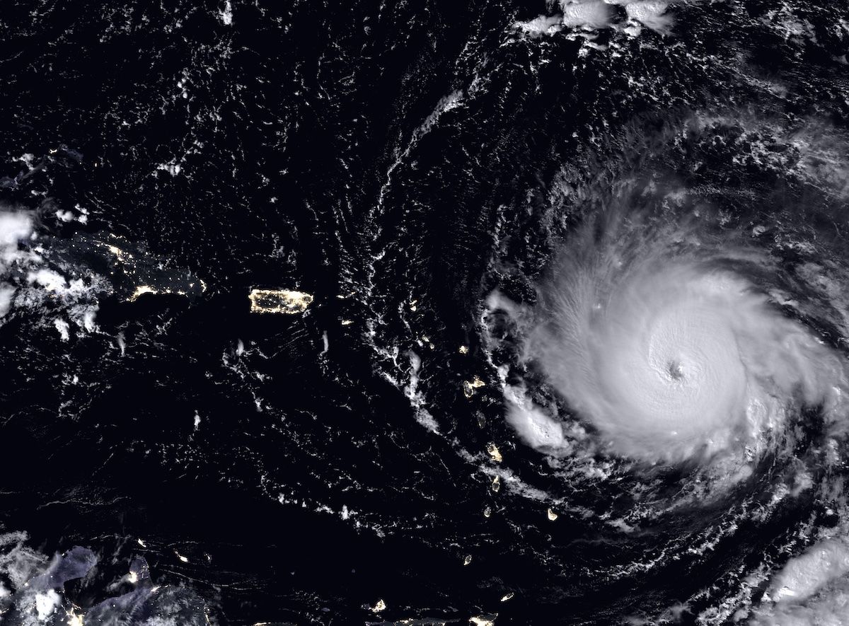 A satellite night image of the Caribbean: Hurricane Irma's vast spiral and clear eye glow at right against black ocean, while the lit outlines of Puerto Rico, Hispaniola and the small Leeward Islands appear as scattered points of light directly in the storm's path.