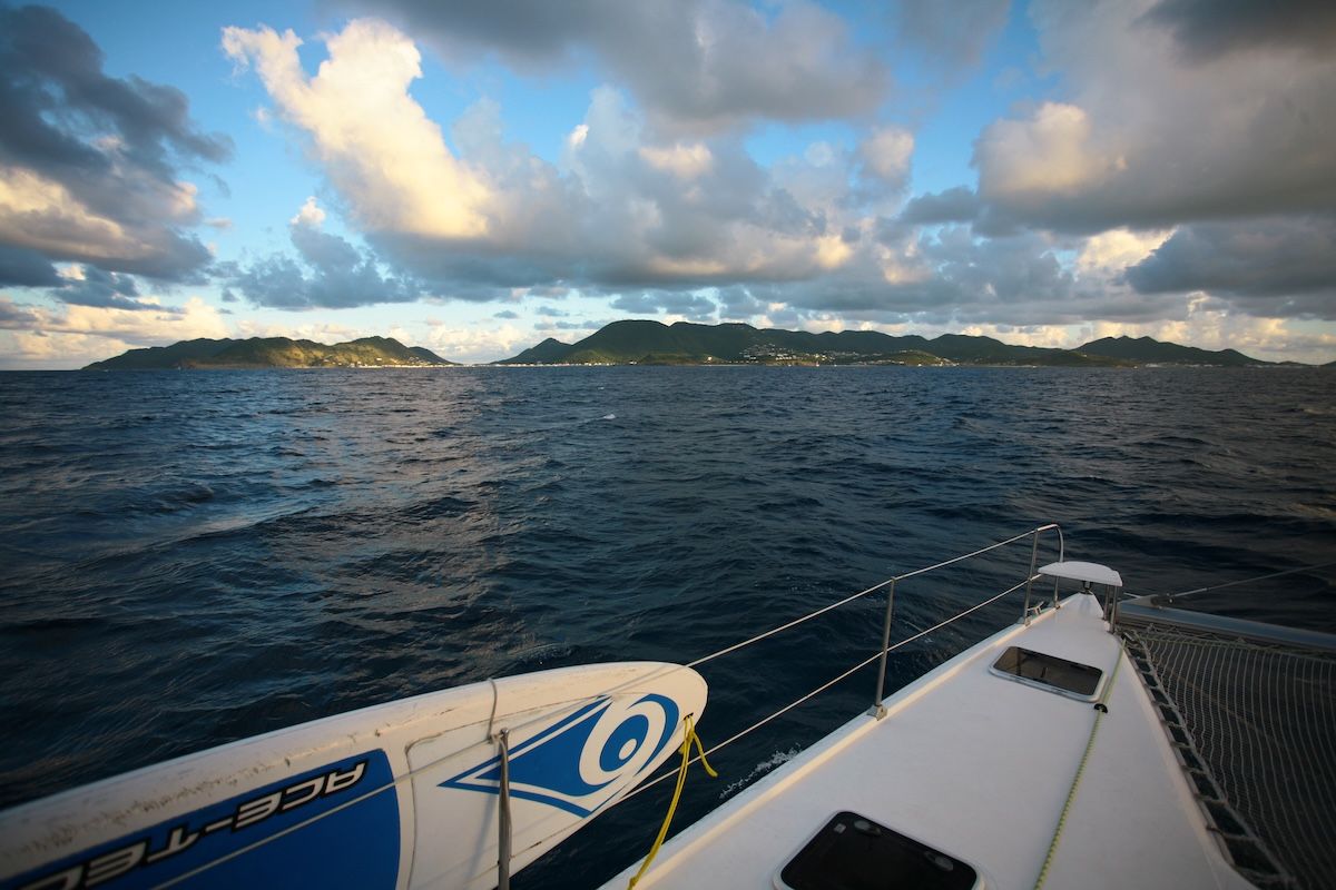 The island seen at dusk from the deck of a sailing boat offshore, its hills running low along the horizon under broken cloud with lights beginning to show along the shore.