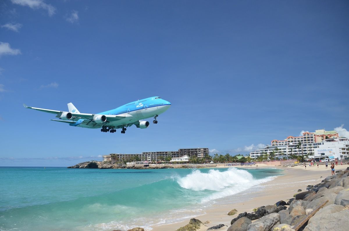 A blue and white KLM Boeing 747 flying very low over Maho Beach on its approach to the runway, with turquoise surf breaking on the sand and hotels along the shoreline below.
