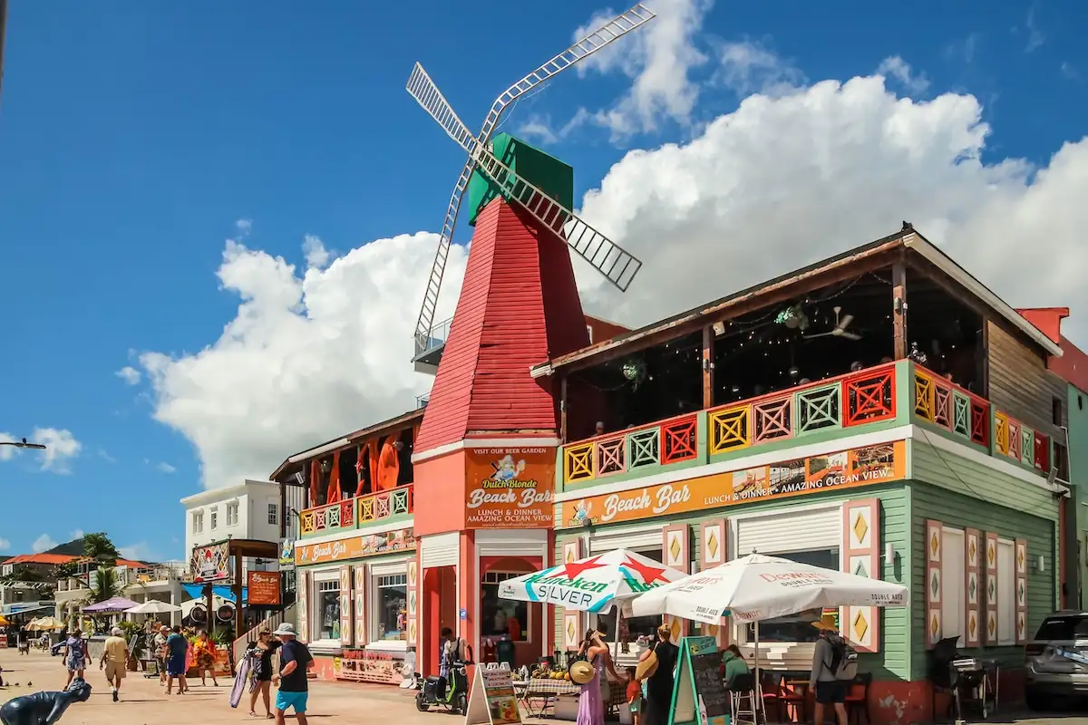 A brightly painted beach bar with a windmill on its roof on the Philipsburg boardwalk, with people walking past under a blue sky