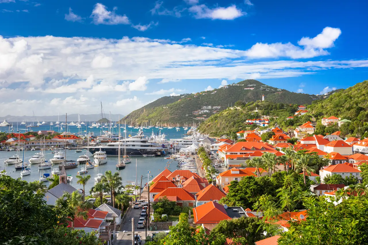 The harbour at Gustavia in daylight, with yachts moored along the quay and red-roofed buildings rising up the hillside behind