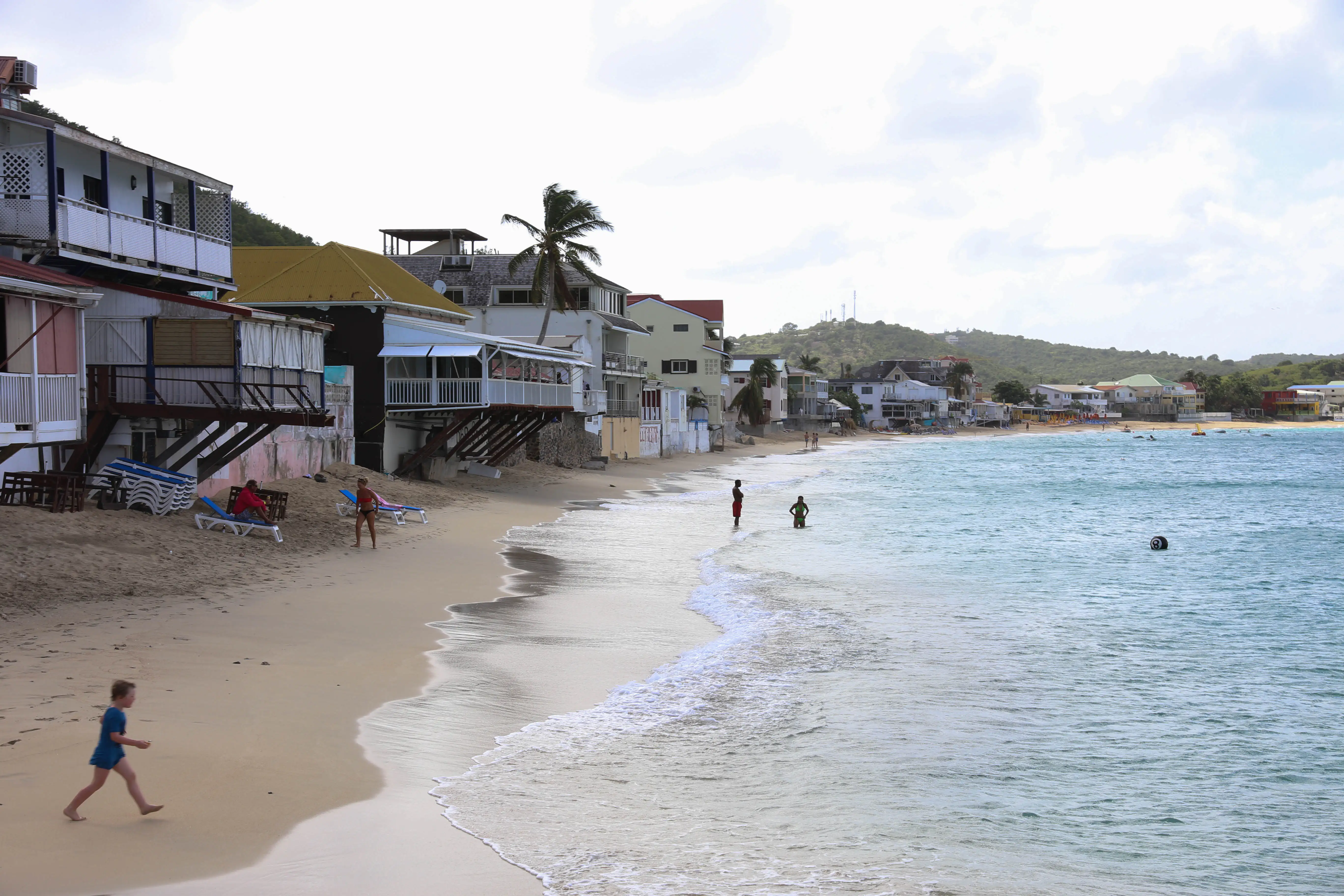 The beach at Grand Case curving away past wooden restaurant buildings raised on stilts at the sand's edge, with loungers, a child walking the waterline and swimmers standing in calm shallow water