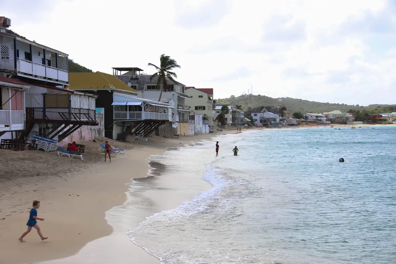 The beach at Grand Case curving away past wooden restaurant buildings raised on stilts at the sand's edge, with loungers, a child walking the waterline and swimmers standing in calm shallow water