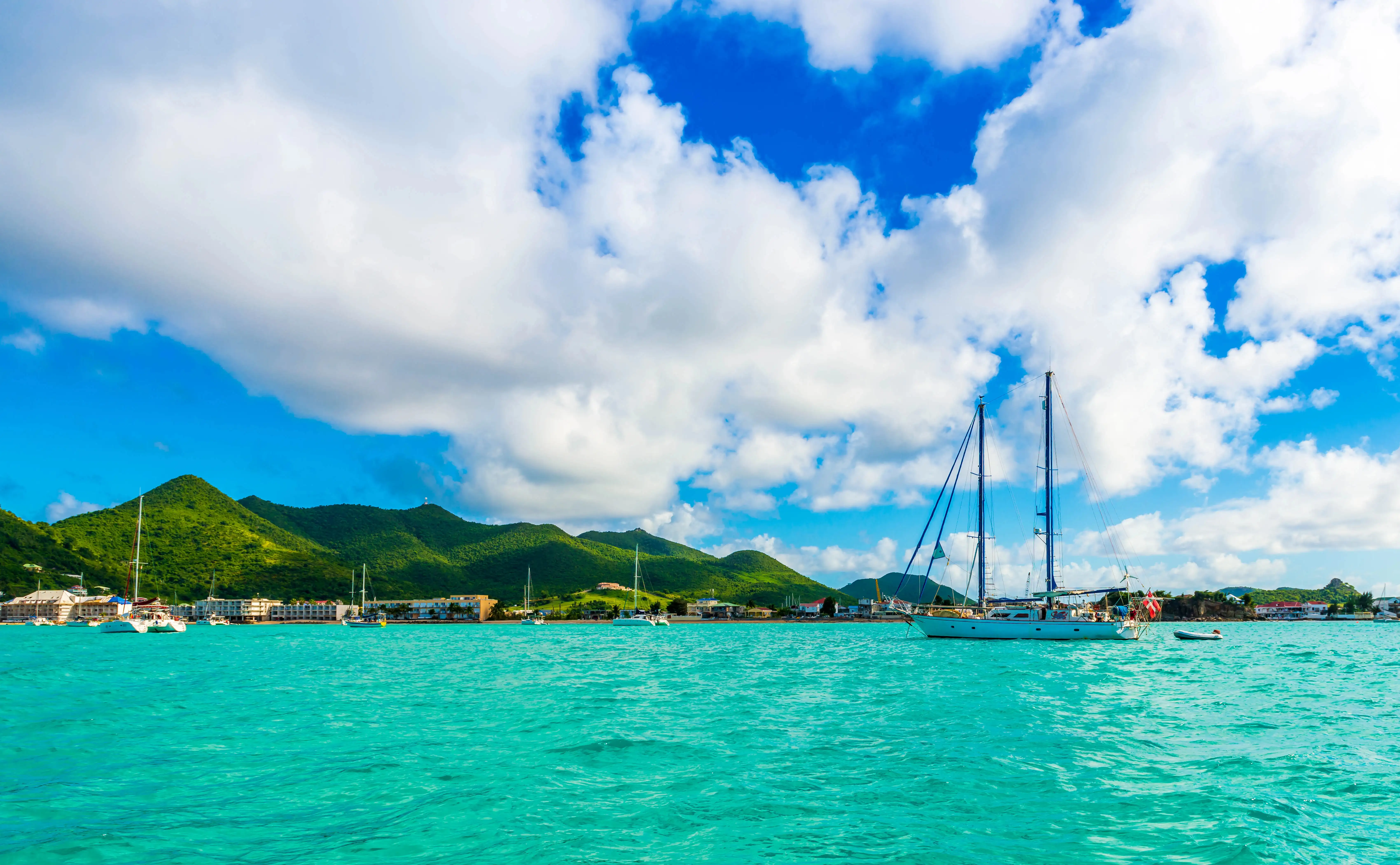 Sailboat anchored in turquoise water off a beachfront row of buildings, with green hills behind