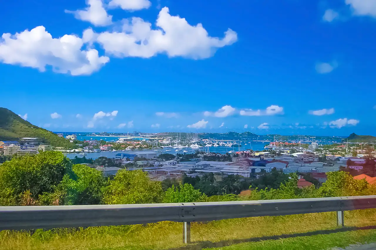 A road running along a hillside above St. Maarten, with a guardrail in the foreground and the town and lagoon spread out below