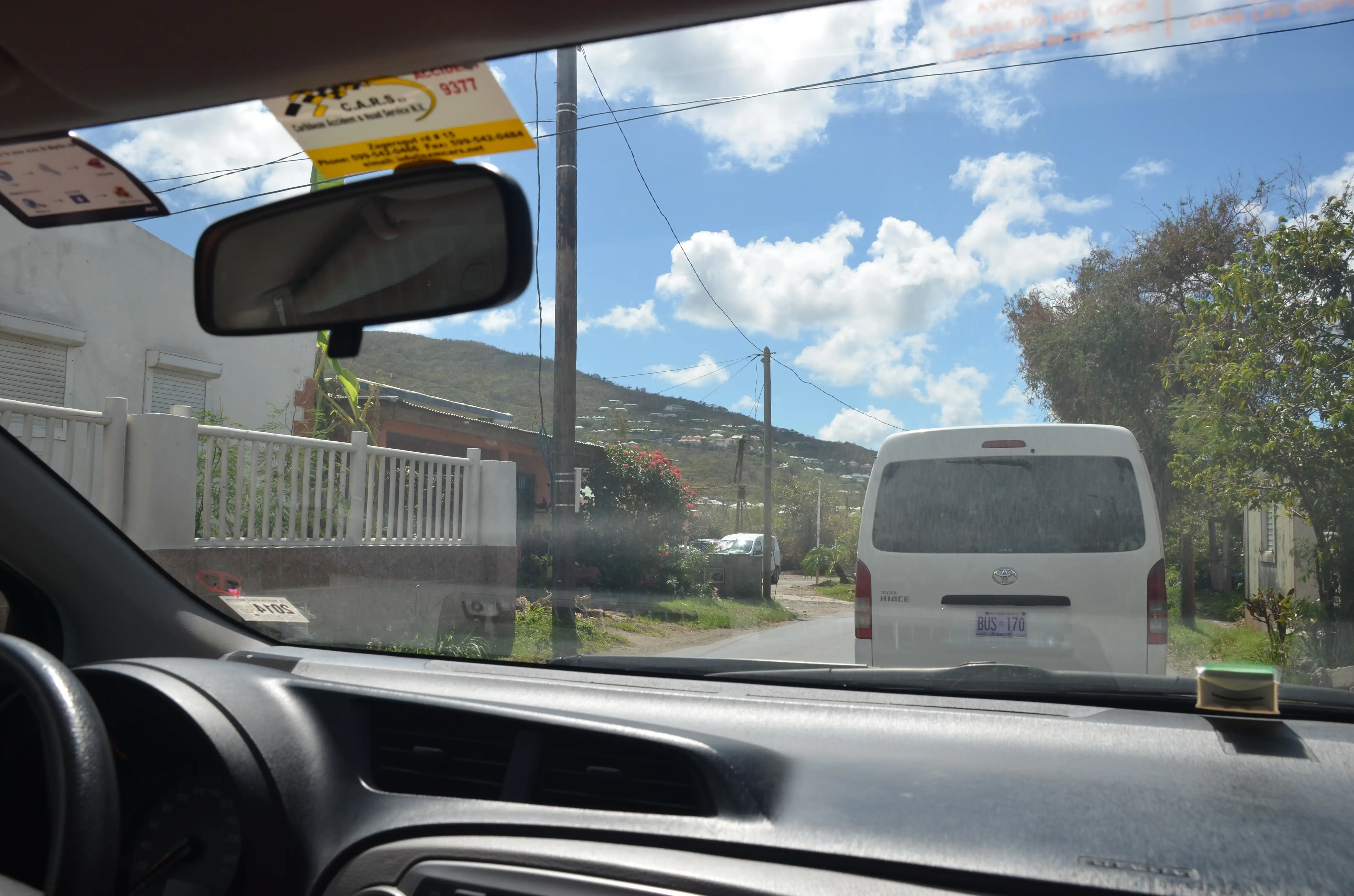 The view through a car windscreen of a queue of traffic behind a white van on a hillside road