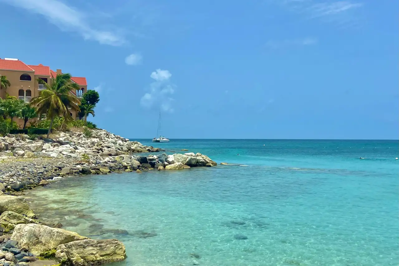 Clear turquoise water washing over a rocky shoreline at Little Bay, with a terracotta-roofed resort building on the headland behind and a catamaran moored offshore