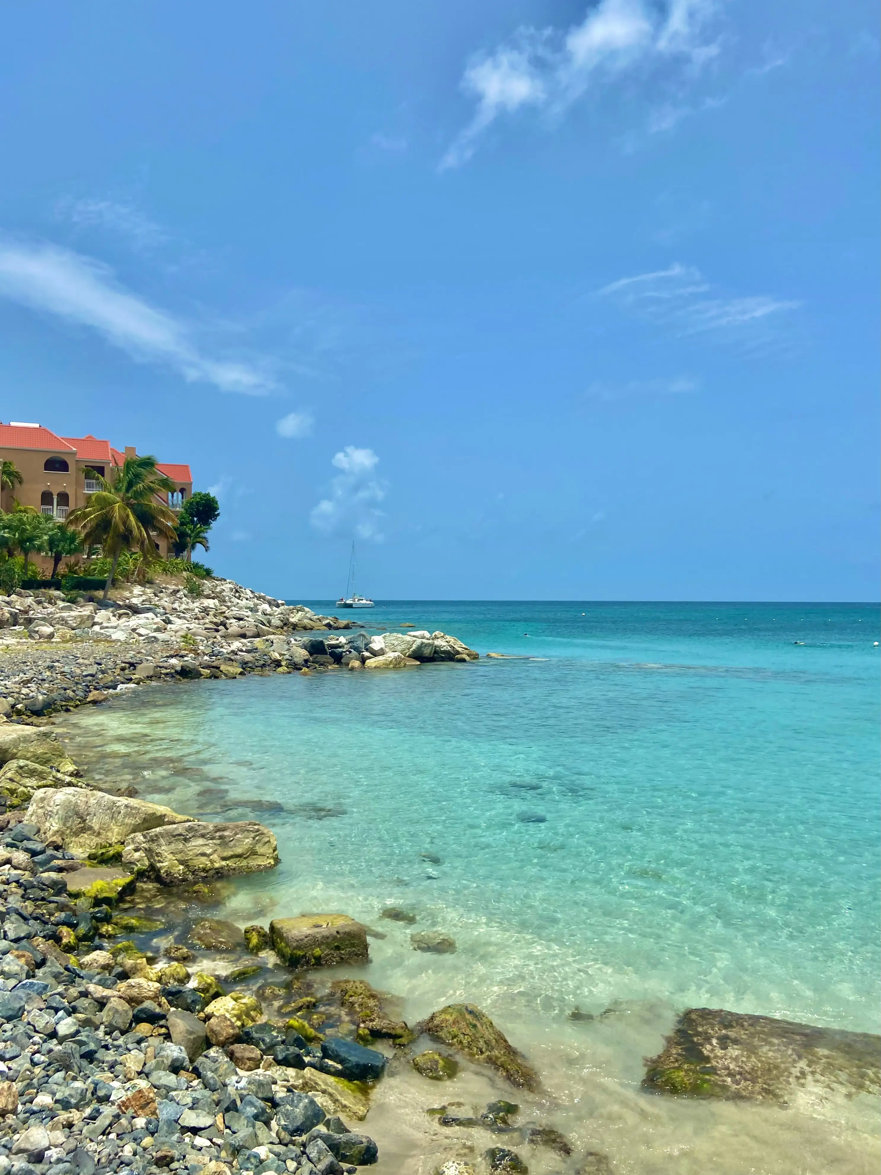 Clear turquoise water washing over a rocky shoreline at Little Bay, with a terracotta-roofed resort building on the headland behind and a catamaran moored offshore