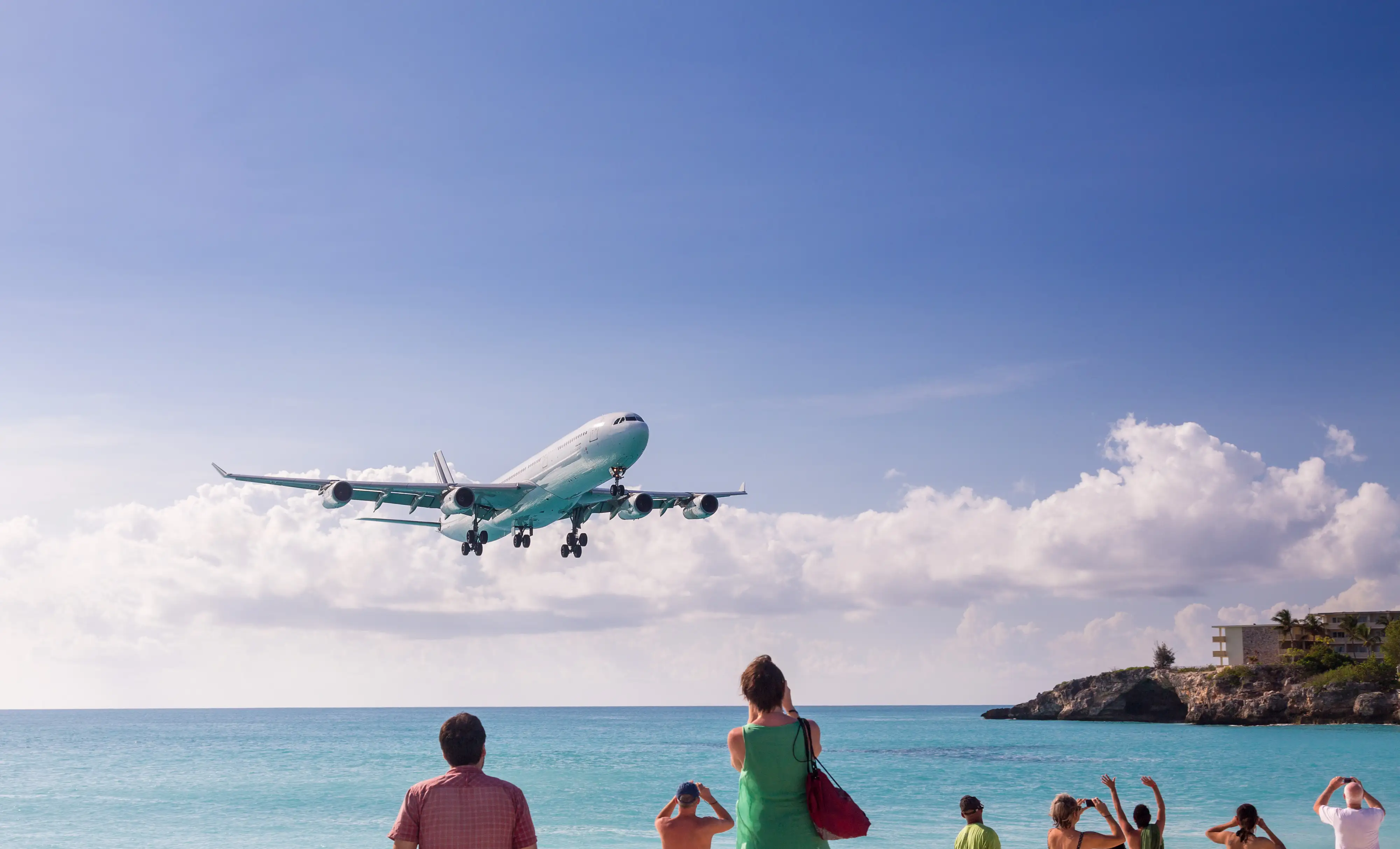 A four engine Airbus A340 on final approach over Maho Beach as a crowd of people photograph it from the sand.