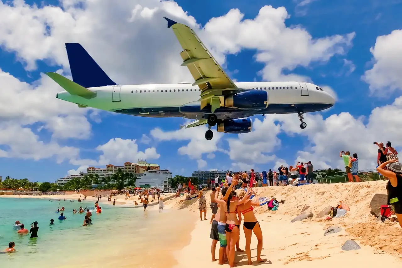 A jet landing low over Maho Beach as beachgoers watch and wave from the sand