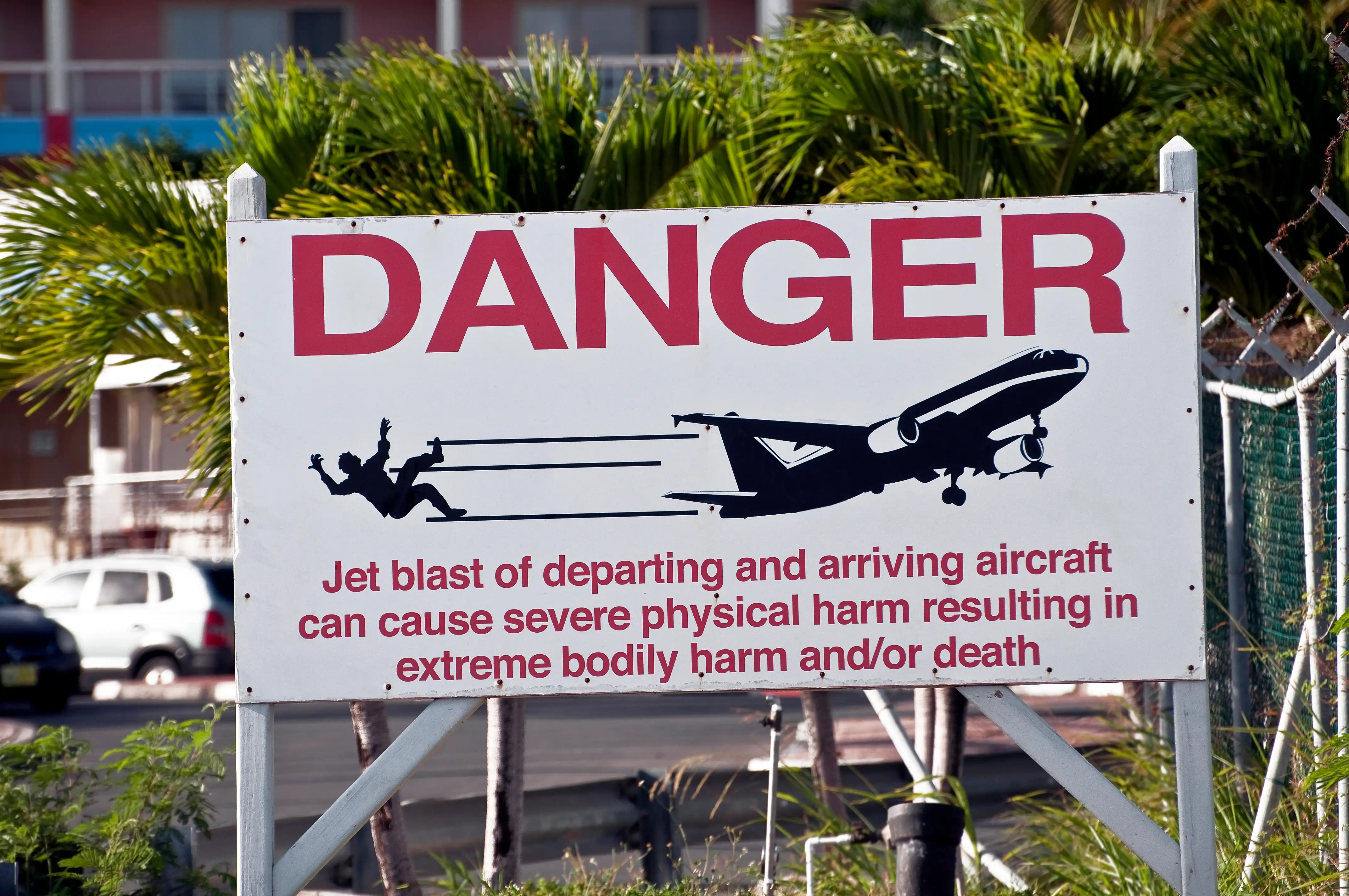 A large warning sign at Maho Beach reading DANGER, with a silhouette of a person being blown off their feet behind a departing jet, and text warning that jet blast can cause severe harm or death.
