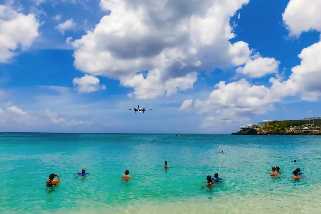 Swimmers in turquoise water at Maho Beach watch a small twin propeller aircraft on final approach, with a cliff and resort buildings on the right.