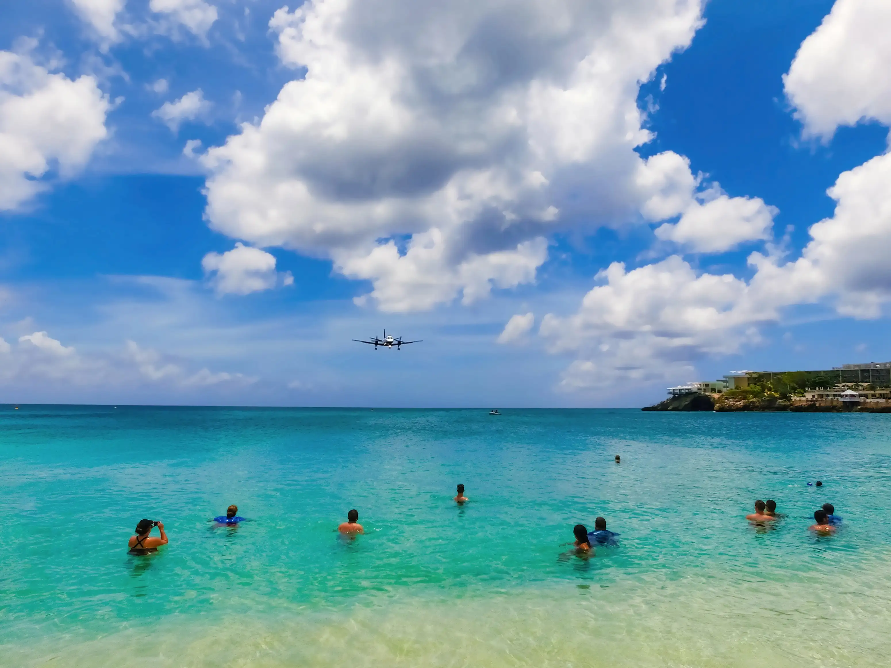 Swimmers in turquoise water at Maho Beach watch a small twin propeller aircraft on final approach, with a cliff and resort buildings on the right.