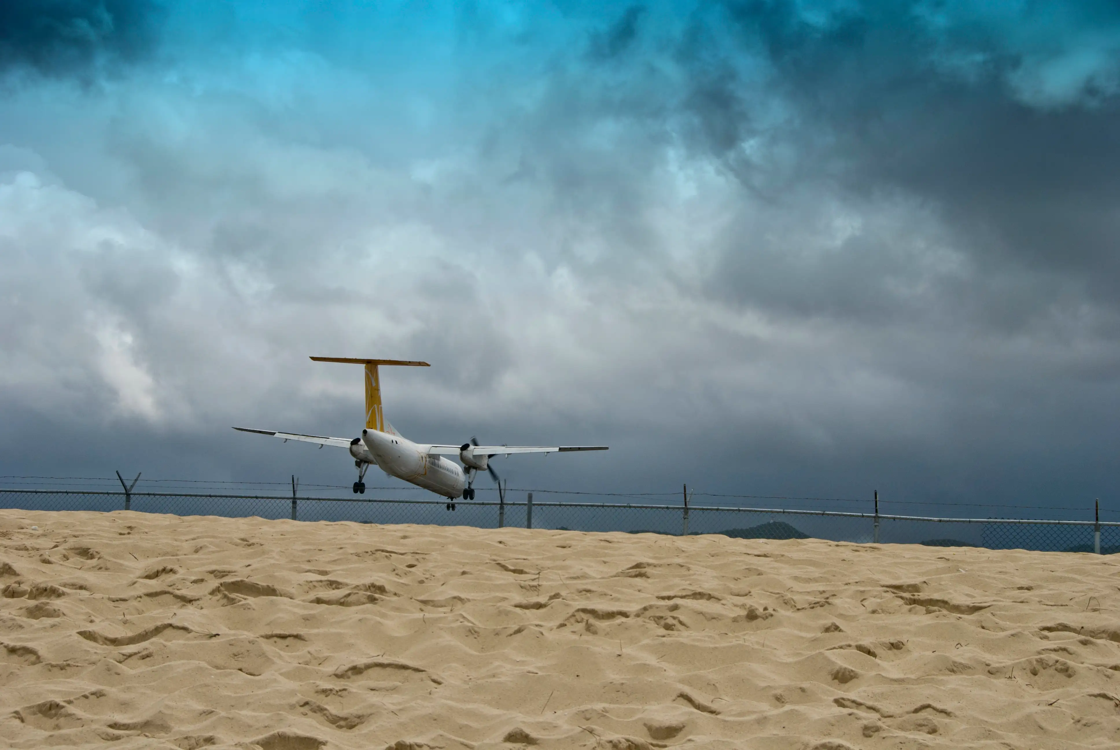 A twin propeller commuter aircraft crosses the airport fence at Maho Beach under a heavy grey sky, seen from the sand.