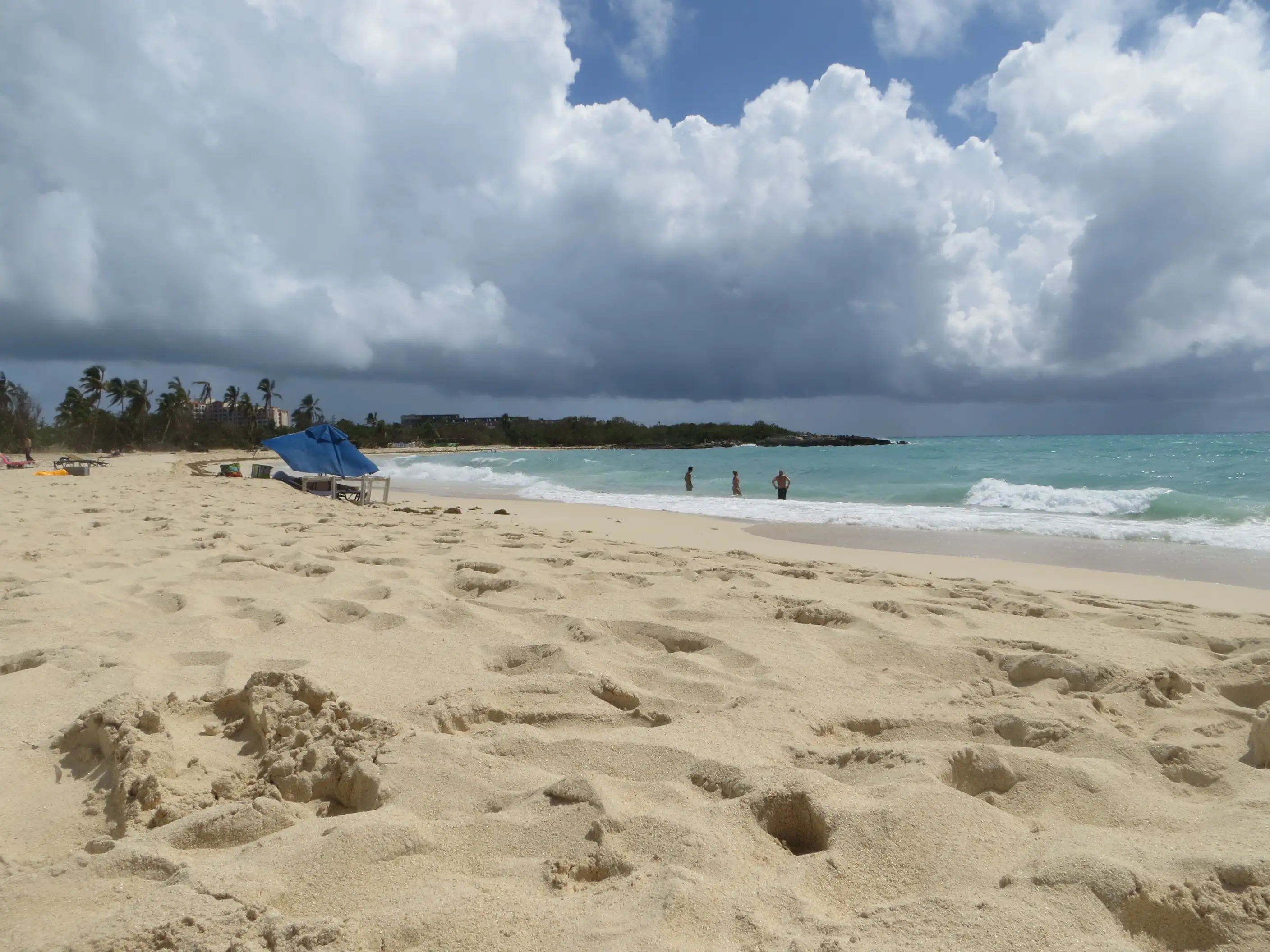 Wide, empty stretch of Mullet Bay Beach under a dramatic cloudy sky, with a few swimmers near the shore