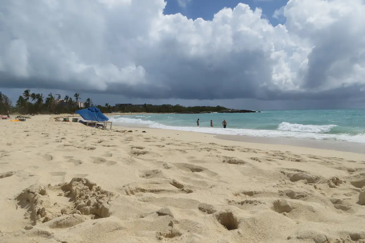 Wide, empty stretch of Mullet Bay Beach under a dramatic cloudy sky, with a few swimmers near the shore