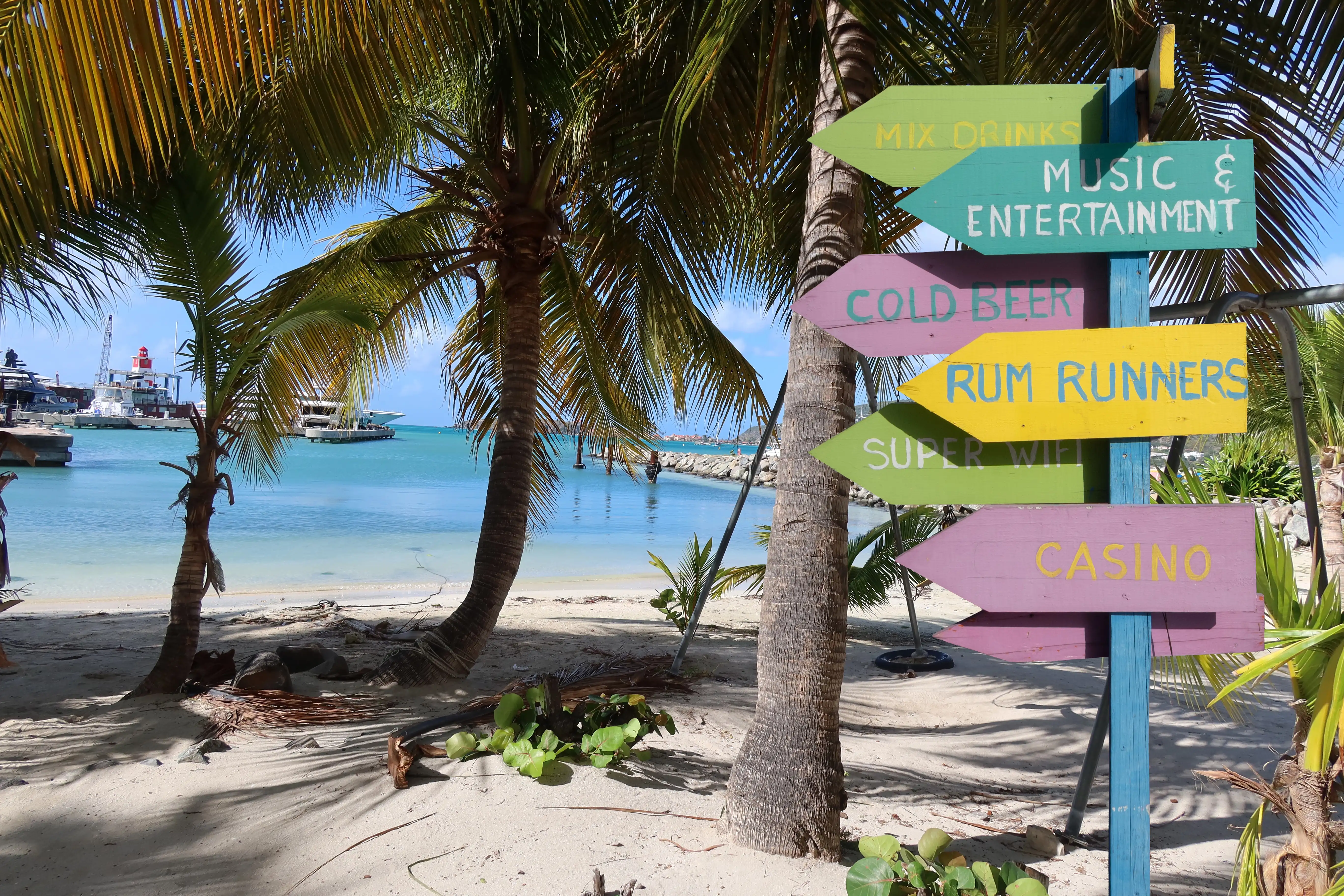 Colorful hand-painted directional signpost on Philipsburg beach pointing to Rum Runners, the casino, and live music, with a cruise ship visible in Great Bay