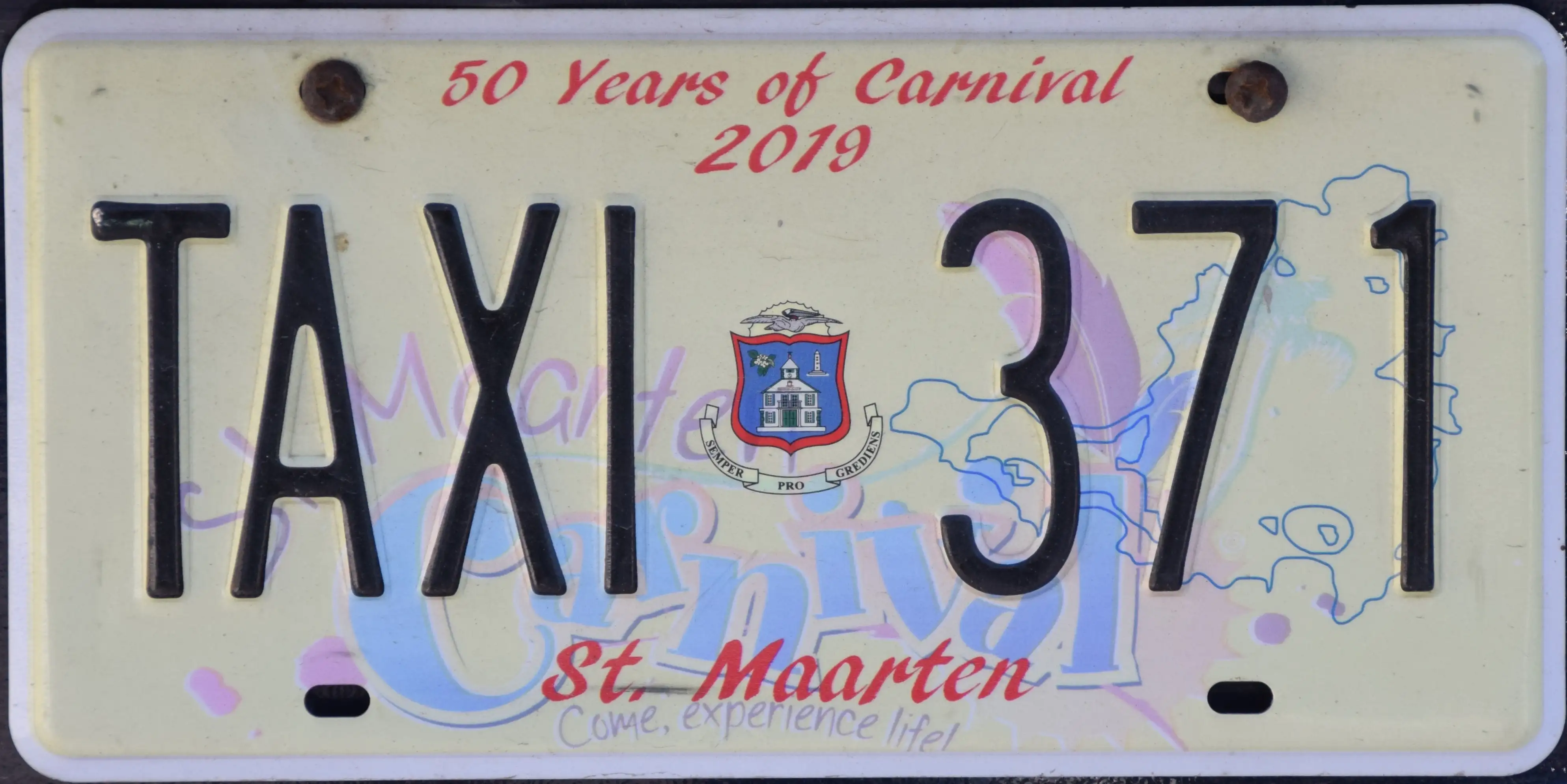 A yellow and white Sint Maarten licence plate reading TAXI 371, mounted on the back of a vehicle above a commemorative Carnival base plate