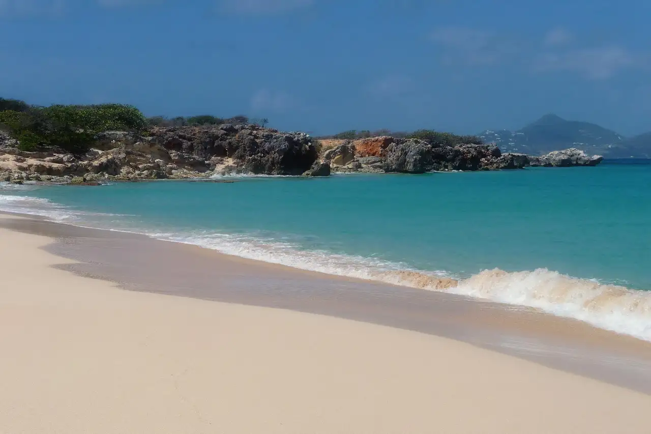 An empty stretch of pale sand on an uninhabited island, with turquoise shallows in front and low dry scrub behind